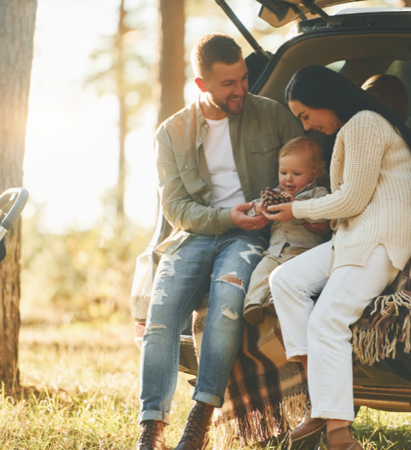 A family of 3 with a mother, father, and toddler, sitting in the back of an SUV in the woods with the sun setting in the background
